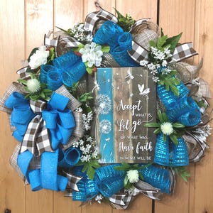 May include: A decorative wreath featuring bright blue mesh loops, brown and white buffalo check ribbon, and white floral accents. A central rectangular sign displays the text: Accept what is, Let go of what was, Have faith in what will be.