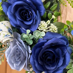Op de afbeelding: Een decoratieve krans met twee grote donkerblauwe stoffen rozen, een kleinere lichtblauwe roos en groen blad. Het arrangement bevat een cr&egrave;mekleurig lint met een blauw bloemenpatroon, kleine witte bloemtrossen en blauwe bessen.
