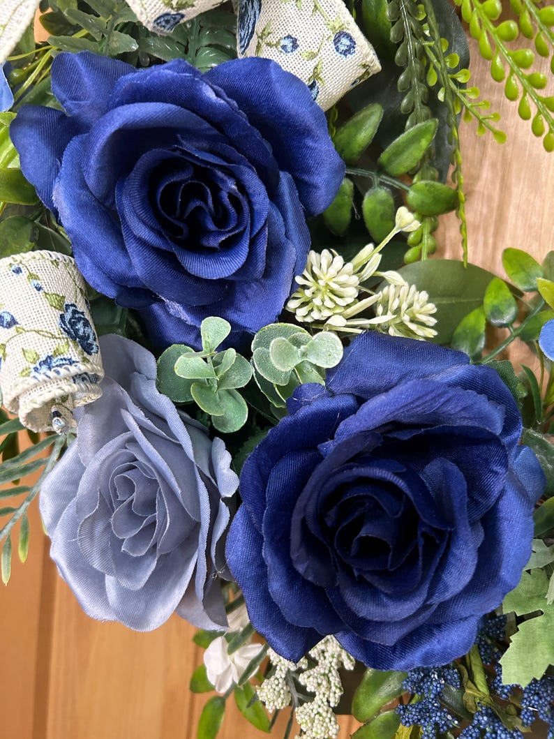 Op de afbeelding: Een decoratieve krans met twee grote donkerblauwe stoffen rozen, een kleinere lichtblauwe roos en groen blad. Het arrangement bevat een cr&egrave;mekleurig lint met een blauw bloemenpatroon, kleine witte bloemtrossen en blauwe bessen.
