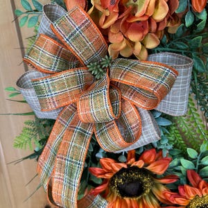 May include: A decorative wreath featuring orange and yellow sunflowers, green foliage, and a large multi-loop bow made of plaid ribbon in shades of orange, green, and beige. The bow is centered among the flowers and greenery, creating a layered arrangement.