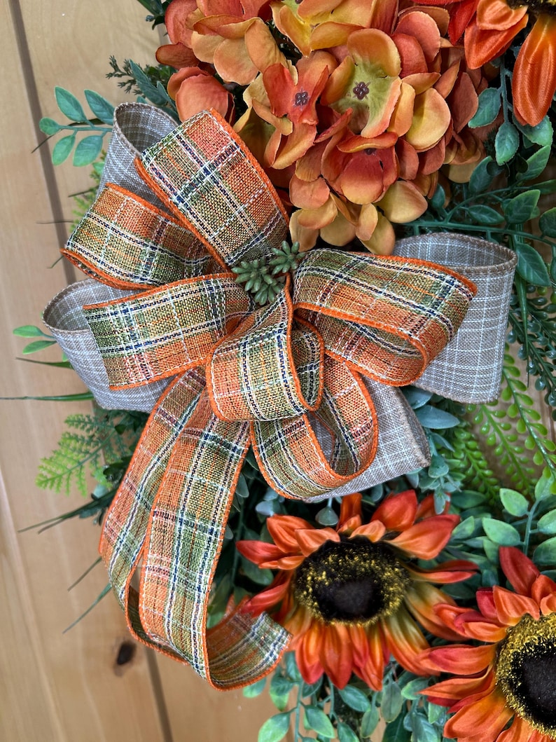 May include: A decorative wreath featuring orange and yellow sunflowers, green foliage, and a large multi-loop bow made of plaid ribbon in shades of orange, green, and beige. The bow is centered among the flowers and greenery, creating a layered arrangement.