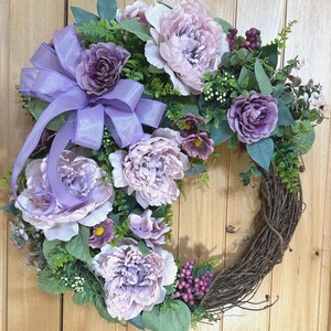 May include: A decorative wreath featuring large light purple and lavender peony flowers, smaller dark purple blossoms, green foliage, and small purple berries. A large, shimmering light purple ribbon bow is attached to the side of the grapevine base.