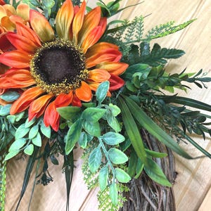 May include: A decorative wreath featuring large, vibrant orange and yellow sunflowers with dark brown centers, accented by various green foliage including fern-like leaves, slender grass blades, and small oval leaves, all attached to a natural twig base.