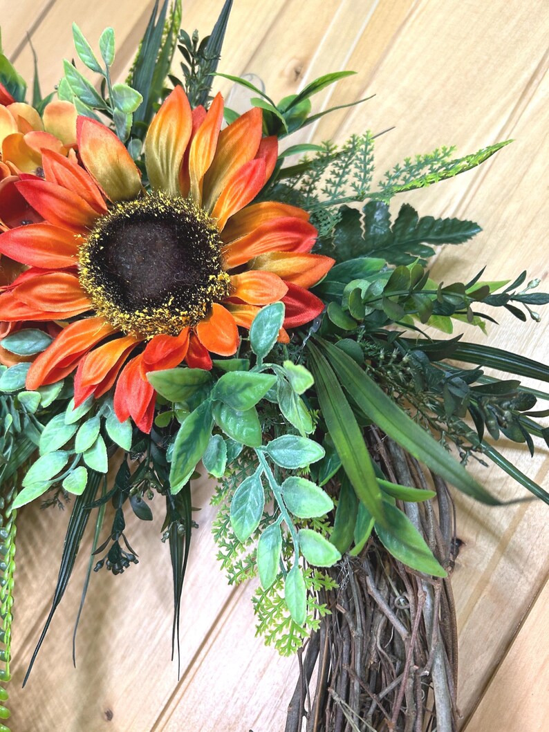 May include: A decorative wreath featuring large, vibrant orange and yellow sunflowers with dark brown centers, accented by various green foliage including fern-like leaves, slender grass blades, and small oval leaves, all attached to a natural twig base.