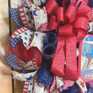 May include: A decorative wreath featuring red, white, and blue mesh ribbons, burlap ribbons with star and stripe patterns, and a large, glittery red bow. A rectangular sign with a star graphic and the partial text AMER is visible on the right.