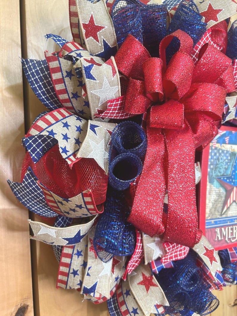 May include: A decorative wreath featuring red, white, and blue mesh ribbons, burlap ribbons with star and stripe patterns, and a large, glittery red bow. A rectangular sign with a star graphic and the partial text AMER is visible on the right.