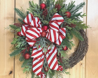 May include: A holiday wreath featuring a natural grapevine base adorned with green pine and cedar branches, dusted with faux snow. The arrangement includes red glittered berry sprigs, small red ornaments, and a large red and white striped ribbon bow.