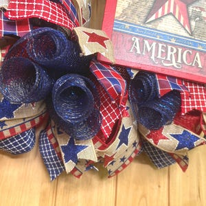May include: A patriotic wreath featuring red, white, and blue mesh rolls and ribbons with star patterns. A rectangular wooden sign with a red border displays a star graphic and the word AMERICA in white lettering, integrated into the design.