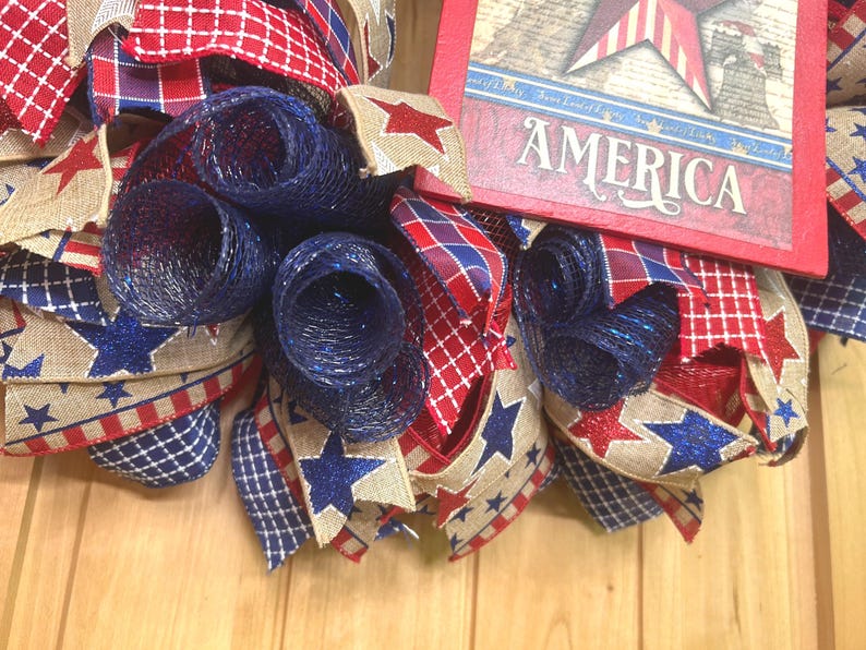 May include: A patriotic wreath featuring red, white, and blue mesh rolls and ribbons with star patterns. A rectangular wooden sign with a red border displays a star graphic and the word AMERICA in white lettering, integrated into the design.