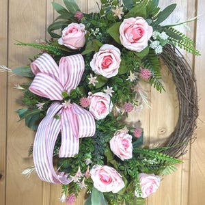 May include: A decorative wreath featuring a natural brown twig base adorned with artificial light pink roses, green foliage, and small pink floral accents. A large, multi-looped bow made of pink and white striped ribbon is attached to the left side.