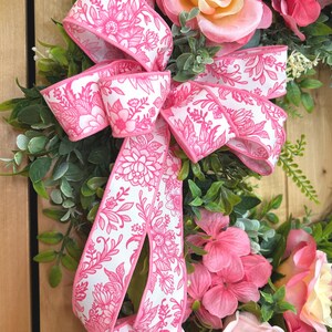May include: A decorative wreath featuring artificial pink and peach roses, green foliage, and a large white ribbon with a vibrant pink floral pattern. The arrangement includes layered petals and lush leaves, creating a full, rounded floral display.
