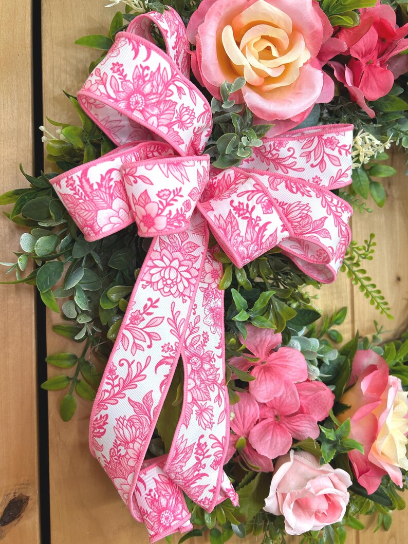 May include: A decorative wreath featuring artificial pink and peach roses, green foliage, and a large white ribbon with a vibrant pink floral pattern. The arrangement includes layered petals and lush leaves, creating a full, rounded floral display.