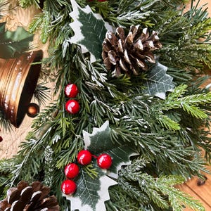 May include: A decorative wreath featuring green artificial pine needles with a light dusting of white snow. The arrangement includes clusters of bright red berries, brown pinecones, and green holly leaves, accented by a gold-toned bell illustration.