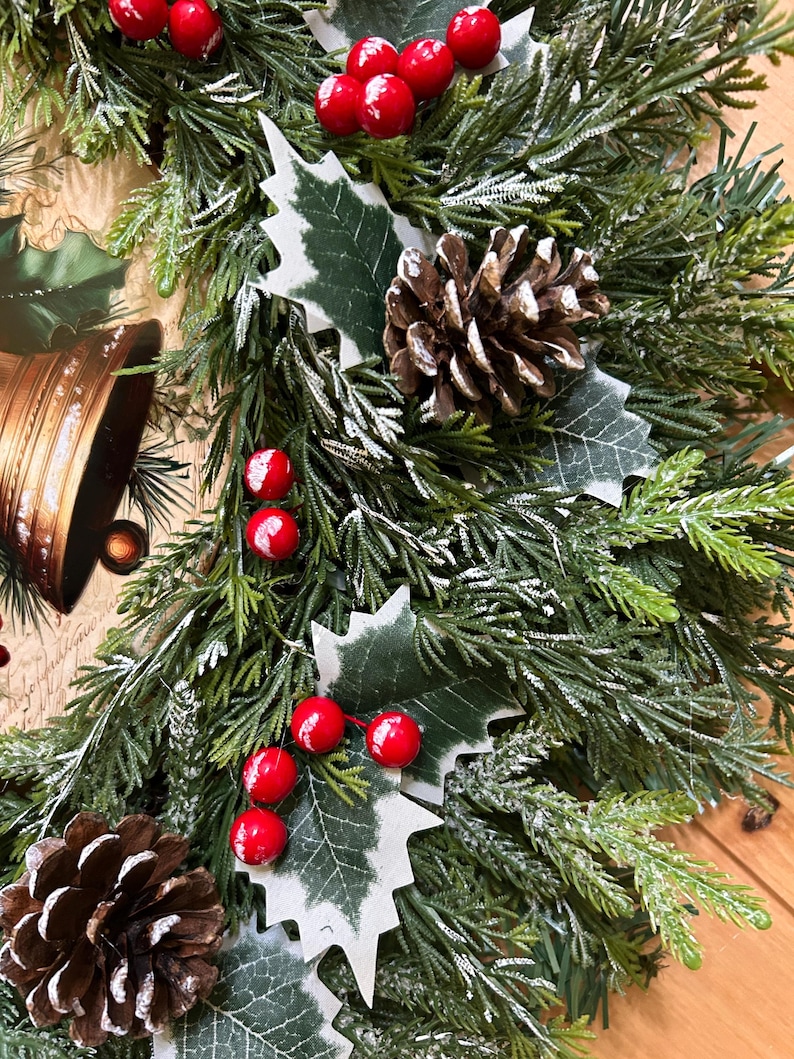 May include: A decorative wreath featuring green artificial pine needles with a light dusting of white snow. The arrangement includes clusters of bright red berries, brown pinecones, and green holly leaves, accented by a gold-toned bell illustration.