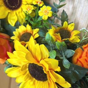 May include: A floral arrangement featuring large yellow sunflowers with dark brown centers, smaller yellow blossoms, and orange flower buds. The display includes green foliage with serrated leaves, creating a dense cluster of artificial botanical elements.