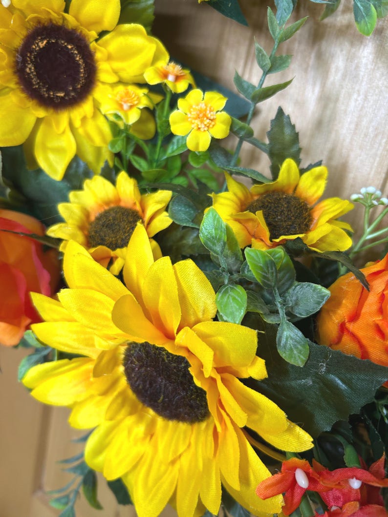 May include: A floral arrangement featuring large yellow sunflowers with dark brown centers, smaller yellow blossoms, and orange flower buds. The display includes green foliage with serrated leaves, creating a dense cluster of artificial botanical elements.