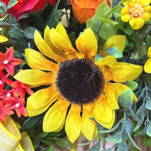May include: A floral arrangement featuring a large yellow sunflower with a dark brown center, surrounded by orange roses, small yellow blossoms, red star-shaped flowers, and green foliage. A yellow and white checkered ribbon is visible at the bottom.
