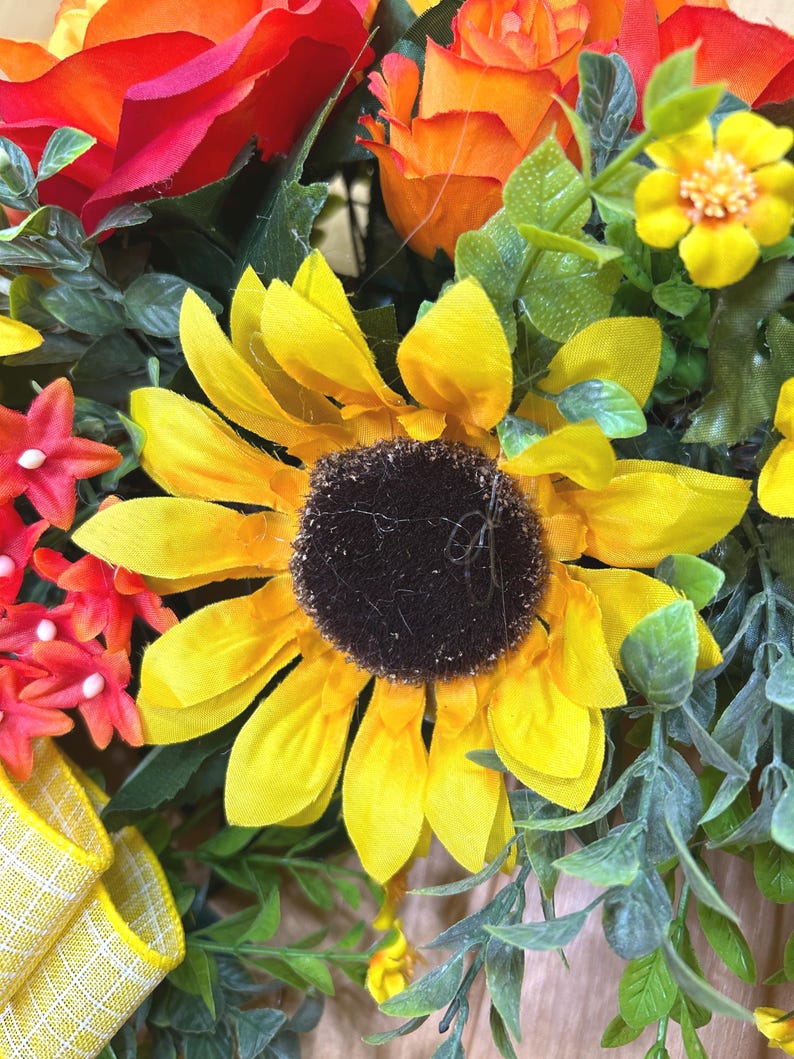 May include: A floral arrangement featuring a large yellow sunflower with a dark brown center, surrounded by orange roses, small yellow blossoms, red star-shaped flowers, and green foliage. A yellow and white checkered ribbon is visible at the bottom.