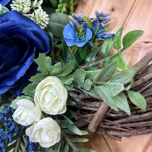 Op de afbeelding: Een decoratieve krans met grote donkerblauwe rozen, kleinere witte rozenknoppen en felblauwe bloesems. Het arrangement bevat groen blad en kleine blauwe bessenclusters, bevestigd op een basis van in elkaar gevlochten bruine takjes.