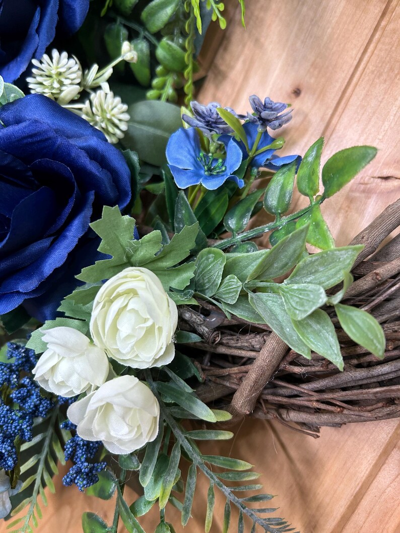 Op de afbeelding: Een decoratieve krans met grote donkerblauwe rozen, kleinere witte rozenknoppen en felblauwe bloesems. Het arrangement bevat groen blad en kleine blauwe bessenclusters, bevestigd op een basis van in elkaar gevlochten bruine takjes.
