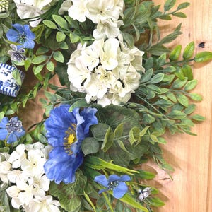 May include: A decorative wreath featuring clusters of white hydrangea-like flowers, vibrant blue poppy-style blossoms, and small blue flowers interspersed with green foliage. A blue and white striped ribbon is visible on the left side of the wreath.