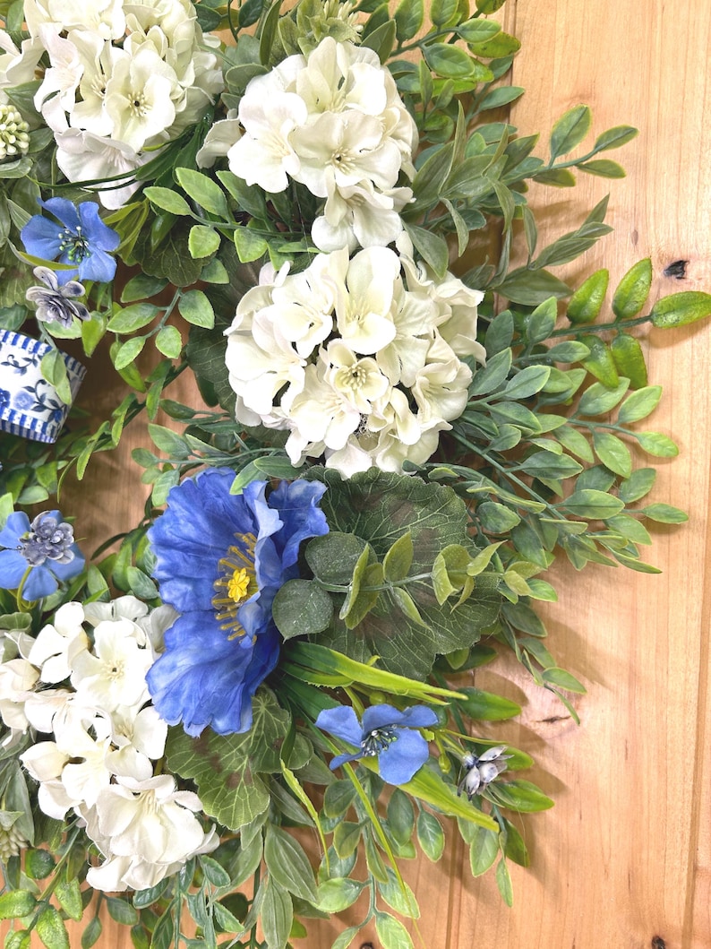 May include: A decorative wreath featuring clusters of white hydrangea-like flowers, vibrant blue poppy-style blossoms, and small blue flowers interspersed with green foliage. A blue and white striped ribbon is visible on the left side of the wreath.