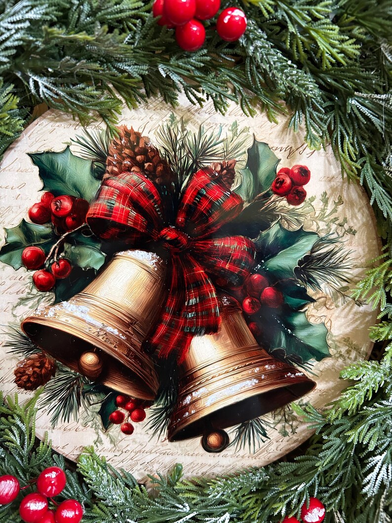 May include: A round decorative plaque featuring a printed illustration of two gold-colored bells tied with a red and green plaid ribbon, surrounded by green holly leaves, red berries, and pine cones against a beige background with script text.