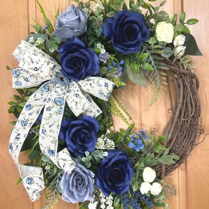 Op de afbeelding: Een decoratieve krans met diep marineblauwe en zachtgrijze rozen, kleine witte bloemen en groen blad op een basis van bruine takken. Een cr&egrave;mekleurig lint met een blauw bloemenpatroon is gestrikt aan de linkerkant van de krans bevestigd.