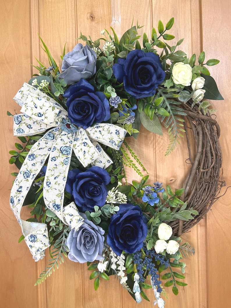 Op de afbeelding: Een decoratieve krans met diep marineblauwe en zachtgrijze rozen, kleine witte bloemen en groen blad op een basis van bruine takken. Een cr&egrave;mekleurig lint met een blauw bloemenpatroon is gestrikt aan de linkerkant van de krans bevestigd.