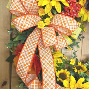 May include: A decorative wreath featuring large yellow sunflowers with dark brown centers, small orange floral clusters, and green foliage. A prominent bow made of white and orange checkered ribbon with orange edges is centered on the arrangement.