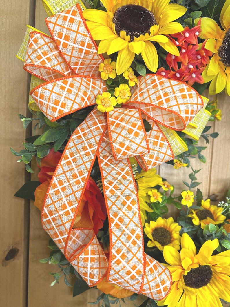 May include: A decorative wreath featuring large yellow sunflowers with dark brown centers, small orange floral clusters, and green foliage. A prominent bow made of white and orange checkered ribbon with orange edges is centered on the arrangement.