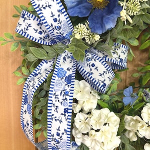 May include: A decorative wreath featuring a large blue flower, clusters of white hydrangea-like blossoms, and green foliage. A white ribbon with a blue floral pattern and blue striped edges is tied in a bow, accenting the arrangement of flowers.