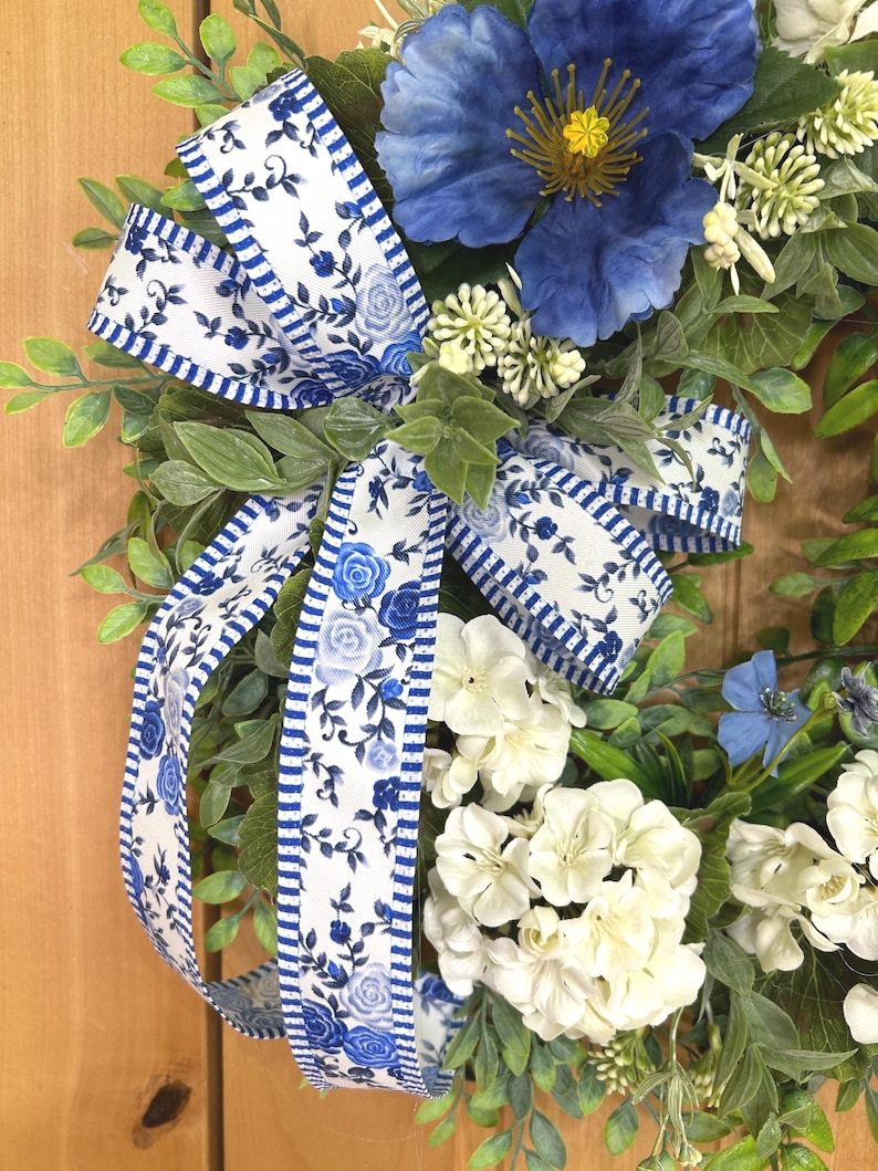 May include: A decorative wreath featuring a large blue flower, clusters of white hydrangea-like blossoms, and green foliage. A white ribbon with a blue floral pattern and blue striped edges is tied in a bow, accenting the arrangement of flowers.