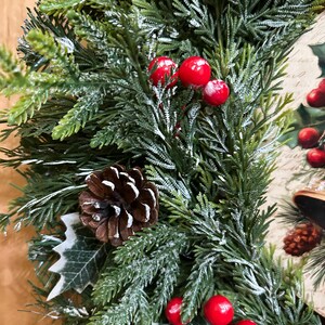 May include: A holiday wreath featuring artificial green pine branches with a light dusting of white snow, accented by clusters of bright red berries, brown pinecones, and green holly leaves, with a decorative printed panel visible on the side.