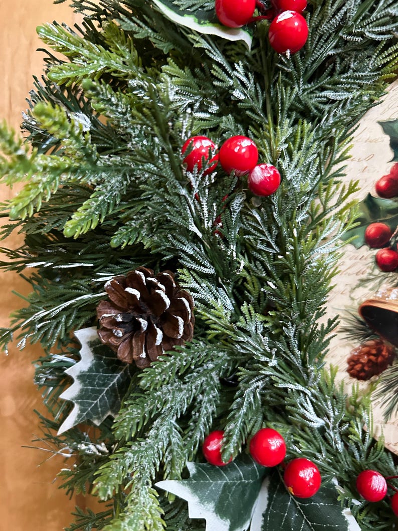 May include: A holiday wreath featuring artificial green pine branches with a light dusting of white snow, accented by clusters of bright red berries, brown pinecones, and green holly leaves, with a decorative printed panel visible on the side.