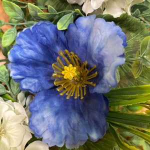 May include: A vibrant blue artificial flower with ruffled petals and a prominent yellow center featuring thin, radiating stamens. The blue flower is surrounded by clusters of white blossoms and green foliage, creating a dense, layered floral arrangement.