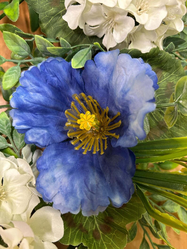 May include: A vibrant blue artificial flower with ruffled petals and a prominent yellow center featuring thin, radiating stamens. The blue flower is surrounded by clusters of white blossoms and green foliage, creating a dense, layered floral arrangement.