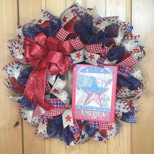 May include: A patriotic wreath featuring red, white, and blue mesh ribbons, burlap ribbons with star patterns, and a large red glittery bow. A rectangular sign in the centre displays a star graphic and the word AMERICA in white lettering.