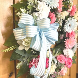 May include: A decorative floral wreath featuring a mix of light blue, white, and pink flowers interspersed with green foliage. A large, prominent bow made of light blue and white striped ribbon is attached to the center, with long tails hanging down.