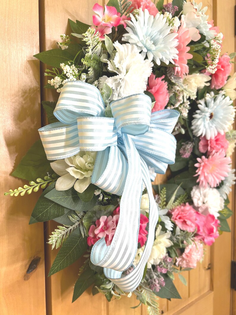 May include: A decorative floral wreath featuring a mix of light blue, white, and pink flowers interspersed with green foliage. A large, prominent bow made of light blue and white striped ribbon is attached to the center, with long tails hanging down.