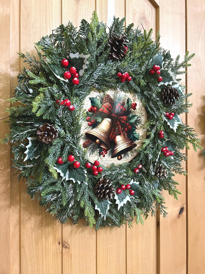 May include: A circular Christmas wreath made of artificial green pine branches, featuring clusters of bright red berries, dark brown pinecones, and white-tipped holly leaves. A central circular plaque displays two gold bells with a red plaid bow.