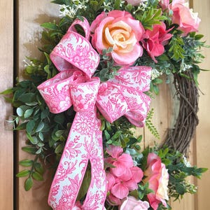 May include: A decorative wreath featuring a brown twig base adorned with green foliage, pink roses, and pink hydrangea clusters. A large, multi-looped ribbon with a white background and a vibrant pink floral line-art pattern is attached to the side.