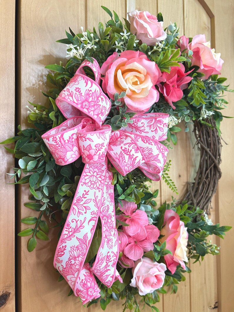 May include: A decorative wreath featuring a brown twig base adorned with green foliage, pink roses, and pink hydrangea clusters. A large, multi-looped ribbon with a white background and a vibrant pink floral line-art pattern is attached to the side.