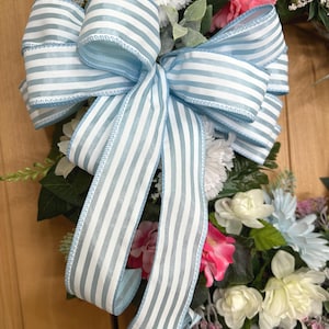 May include: A decorative wreath featuring a large, multi-looped bow made of light blue and white striped ribbon. The wreath is adorned with an assortment of artificial flowers, including white and pink carnations, cream-colored blossoms, and green foliage.