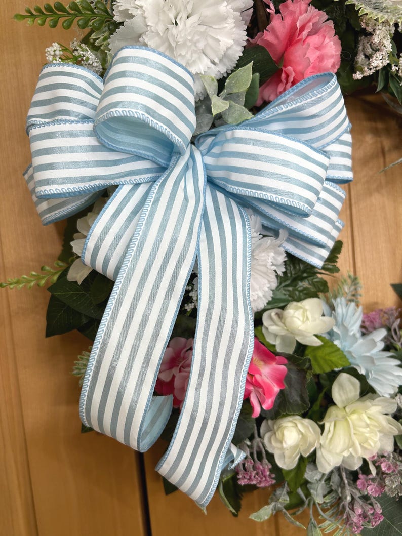 May include: A decorative wreath featuring a large, multi-looped bow made of light blue and white striped ribbon. The wreath is adorned with an assortment of artificial flowers, including white and pink carnations, cream-colored blossoms, and green foliage.