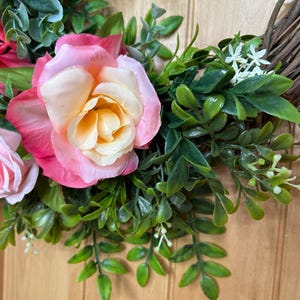May include: A decorative wreath featuring artificial pink and cream roses, vibrant green foliage, and small white flowers attached to a natural brown twig base. The arrangement includes layered rose petals and various green leaves.