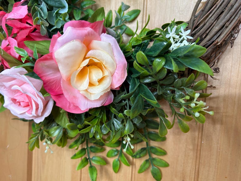 May include: A decorative wreath featuring artificial pink and cream roses, vibrant green foliage, and small white flowers attached to a natural brown twig base. The arrangement includes layered rose petals and various green leaves.