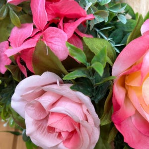 May include: A decorative wreath featuring artificial flowers in shades of bright pink, soft pastel pink, and a blend of pink and yellow. The arrangement includes green foliage and leaves interspersed among the fabric rose and hydrangea blossoms.