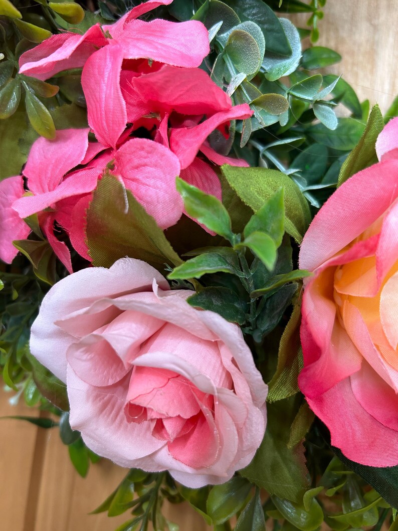 May include: A decorative wreath featuring artificial flowers in shades of bright pink, soft pastel pink, and a blend of pink and yellow. The arrangement includes green foliage and leaves interspersed among the fabric rose and hydrangea blossoms.