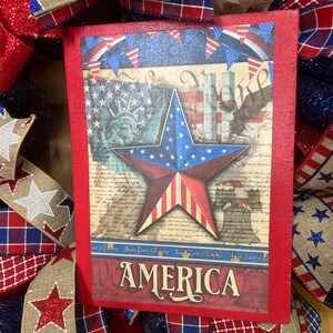 May include: A rectangular wooden sign with a red border featuring a central star with American flag patterns, the Statue of Liberty, a bald eagle, and the Liberty Bell. The text AMERICA is printed at the bottom, surrounded by patriotic ribbon.